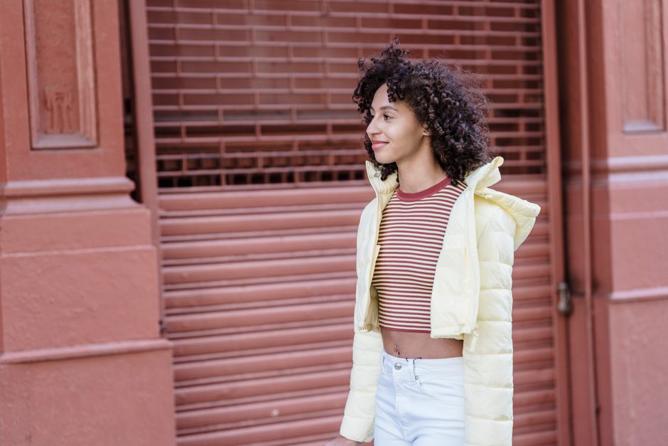 Cheerful ethnic female in padded jacket looking away against city construction with ribbed wall in daytime