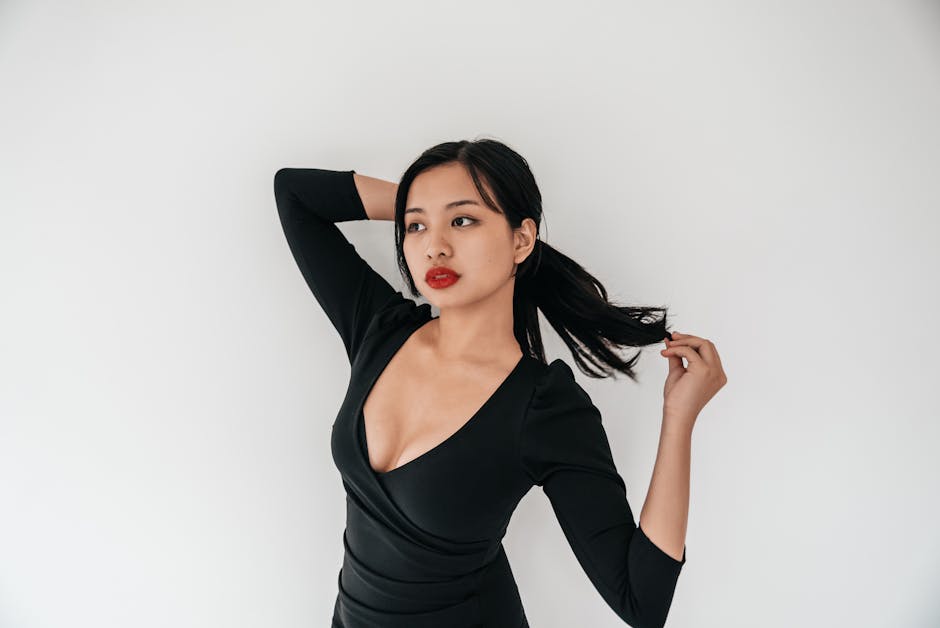 A stylish Asian woman in a black dress poses gracefully indoors against a minimalistic background.