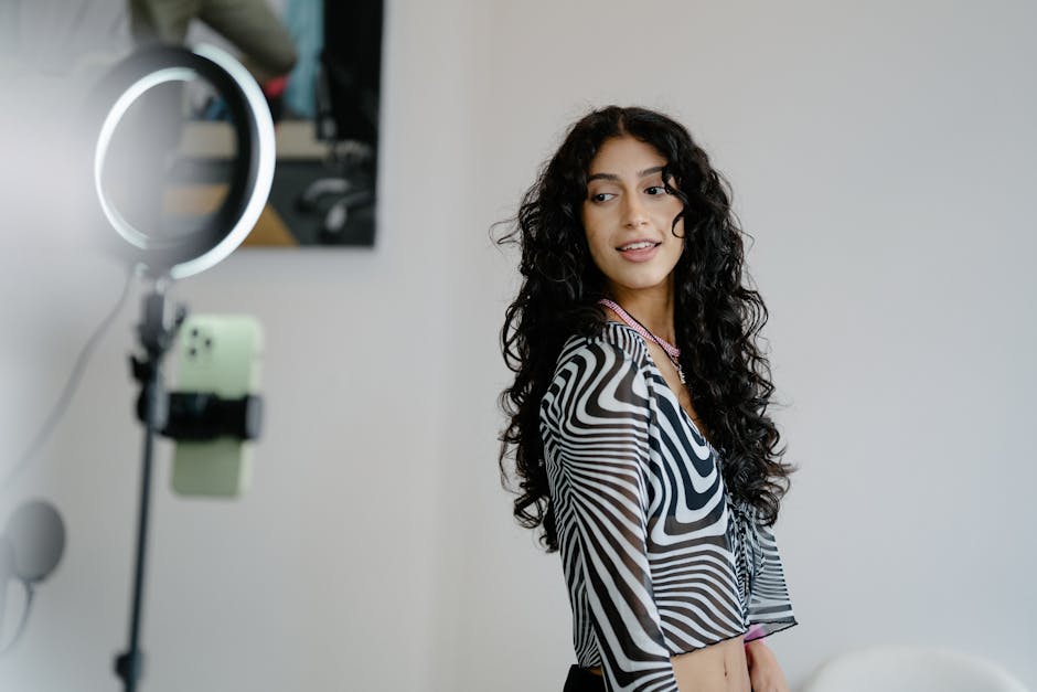 Woman with curly hair using smartphone and ring light for vlogging, wearing a patterned top.