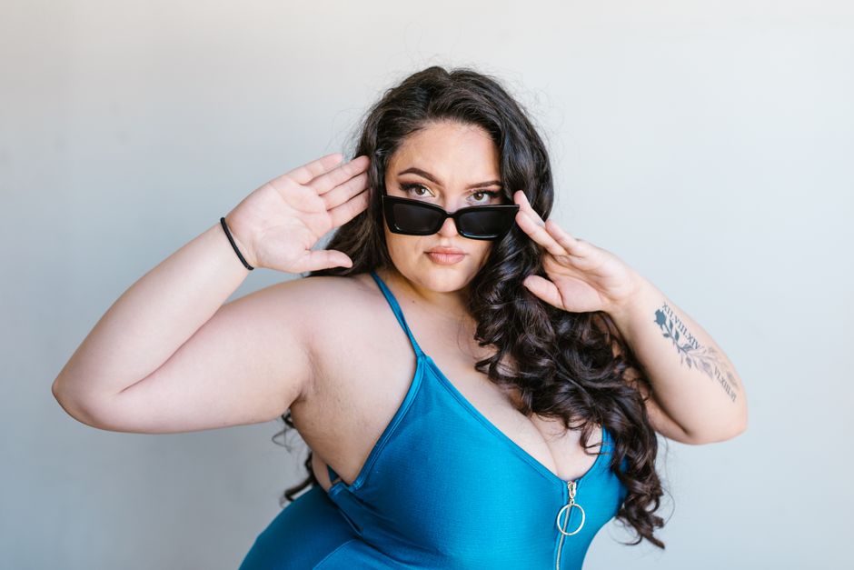 A stylish plus-size woman posing confidently in a blue swimsuit with sunglasses, exuding self-love.