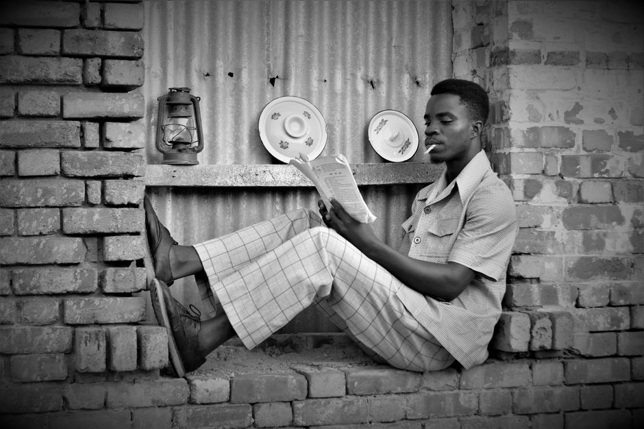 Black and white photo of a man reading and smoking in a relaxed pose against a brick wall.