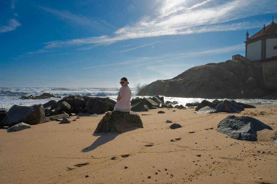 A serene beach with a woman sitting on a rock, waves crashing, and a chapel on the rocky coastline.