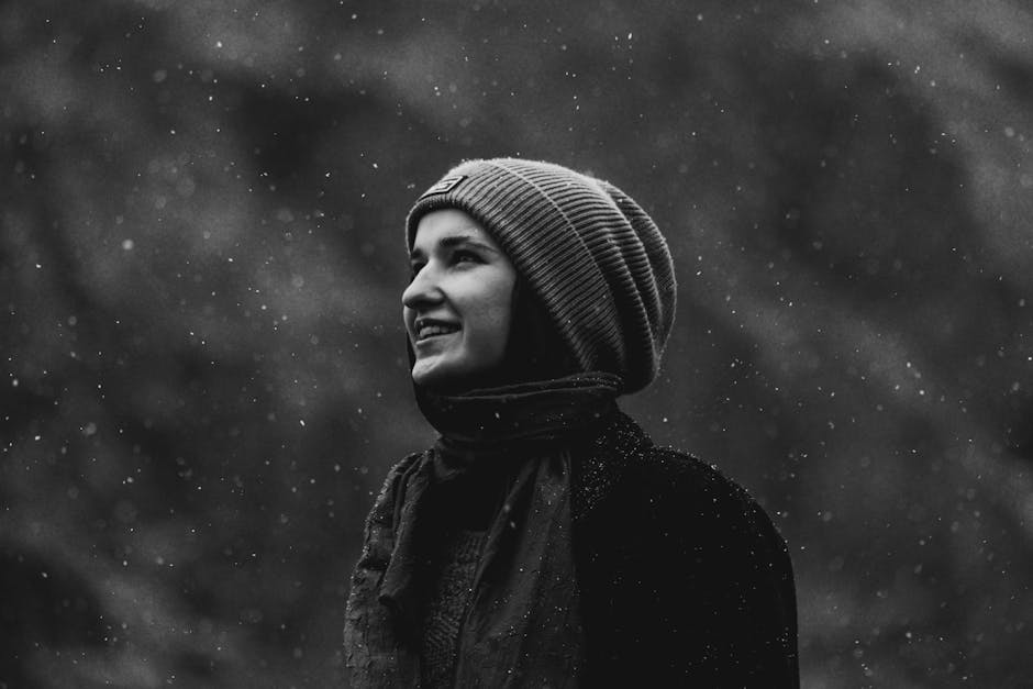 A monochrome portrait of a woman smiling while surrounded by falling snow outdoors.
