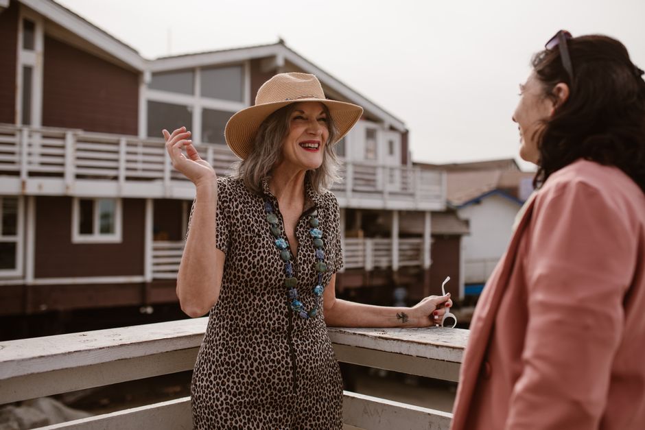 Two women enjoying a conversation on a wooden railing outside on a sunny day.